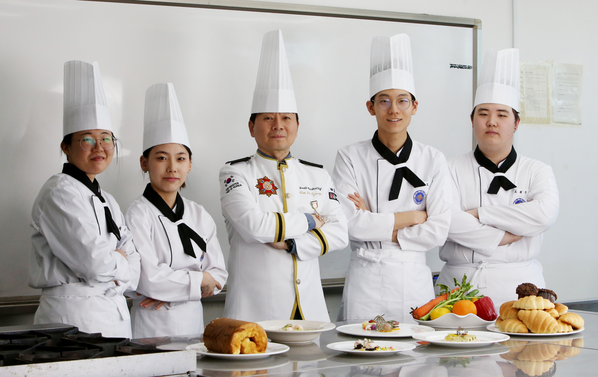 조리실에서 조리복을 입고 조리된 음식 앞에서 팔짱을 끼고 카메라를 바라보는 학생들과 교수님의 사진