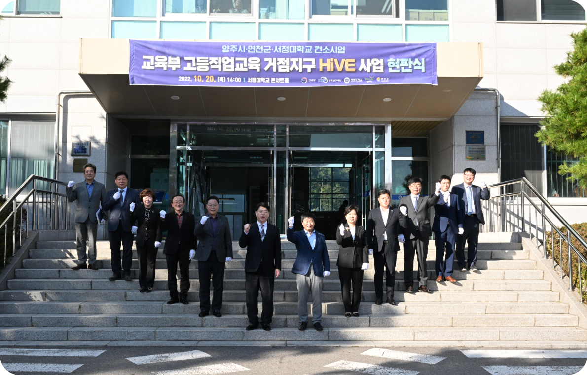 '양주시 연천군 서정대학교 컨소시업, 교육부 거저지구 하이브 사업 현판식'이라고 적힌 플랜카드가 적힌 하이브센터의 정문 사진. 직원 또는 관계자로 보이는 12명의 사람들이 정문 앞 계단에 브이자로 서서 한 손 주먹 쥐어 화이팅 포즈를 하고 있다.