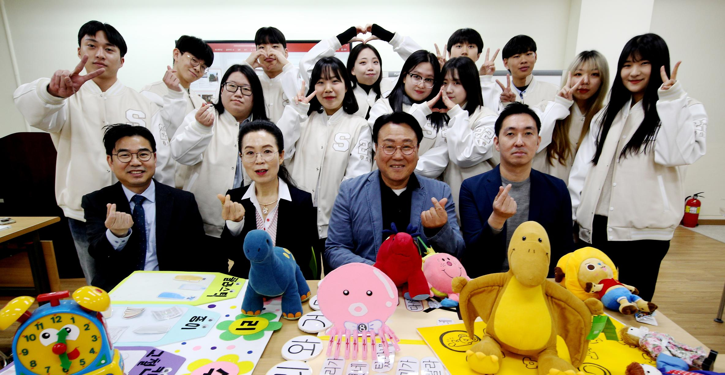 손가락 하트를 하며 책상 앞에 앉은 학생들과 그 뒤에 서있는 교수님들 사진