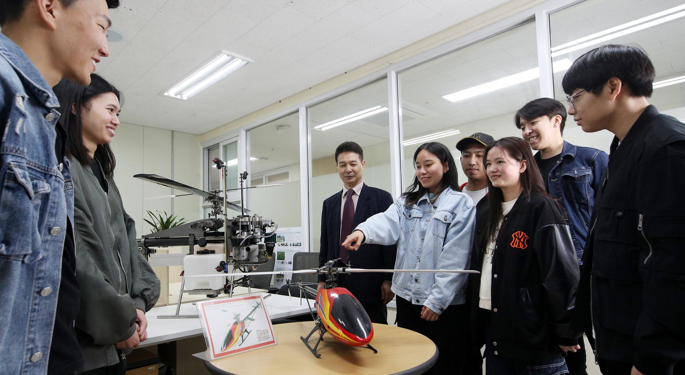 모형 헬리콥터를 사이에 두고 둥글게 모여 토론중인 학생들(7명 이상)과 정장을 입은 교수님 사진