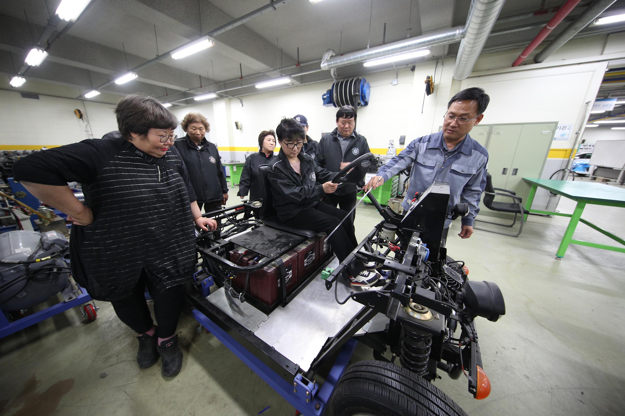 윗부분이 분해 되어 부품이 드러난 자동차 위에 앉아있는 교수님 두 분과 학생 한 명, 그 뒤에 일렬로 서있는 학생들이 한 손 주먹을 쥐고 들어보이는 포즈를 취하고 있다.