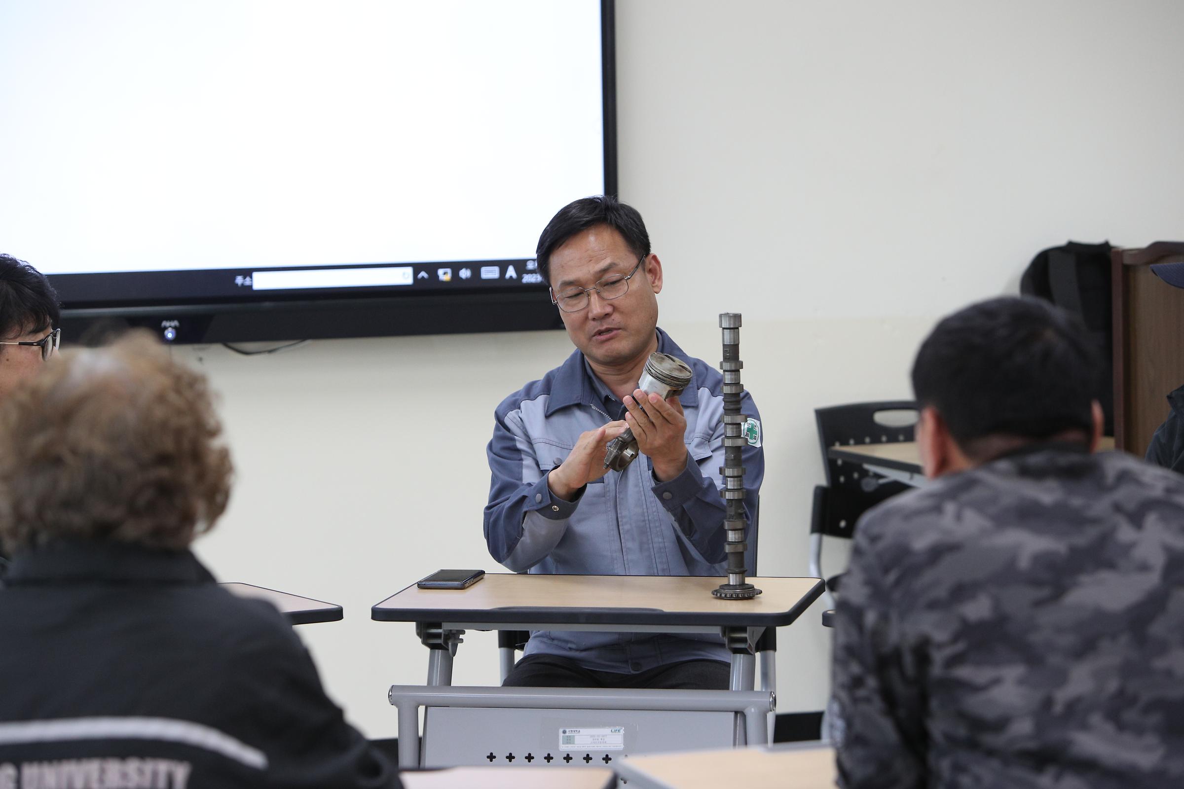 기계 위에 올라가 있는 자동차와 그 자동차와 연경 된 컴퓨터 앞에서 열정적으로 설명 하는 교수님과 경청하는 세명의 학생들