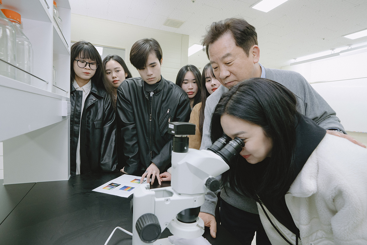 실습실에서 한 학생이 현미경으로 보고있고, 교수님은 뒤에서 다른 학생들은 옆에서 보고 있다.